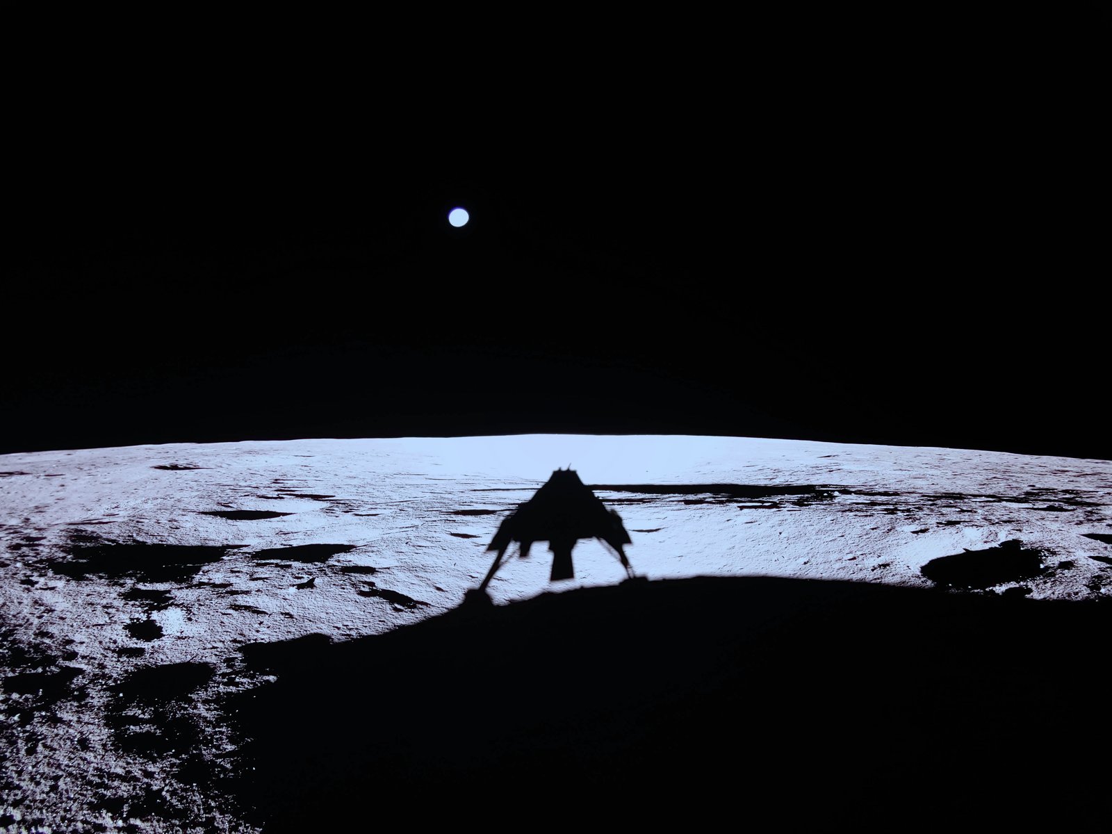 Shadow of a squat four-legged lander on sunlit lunar regolith, Earth as a small blue disc in a black sky