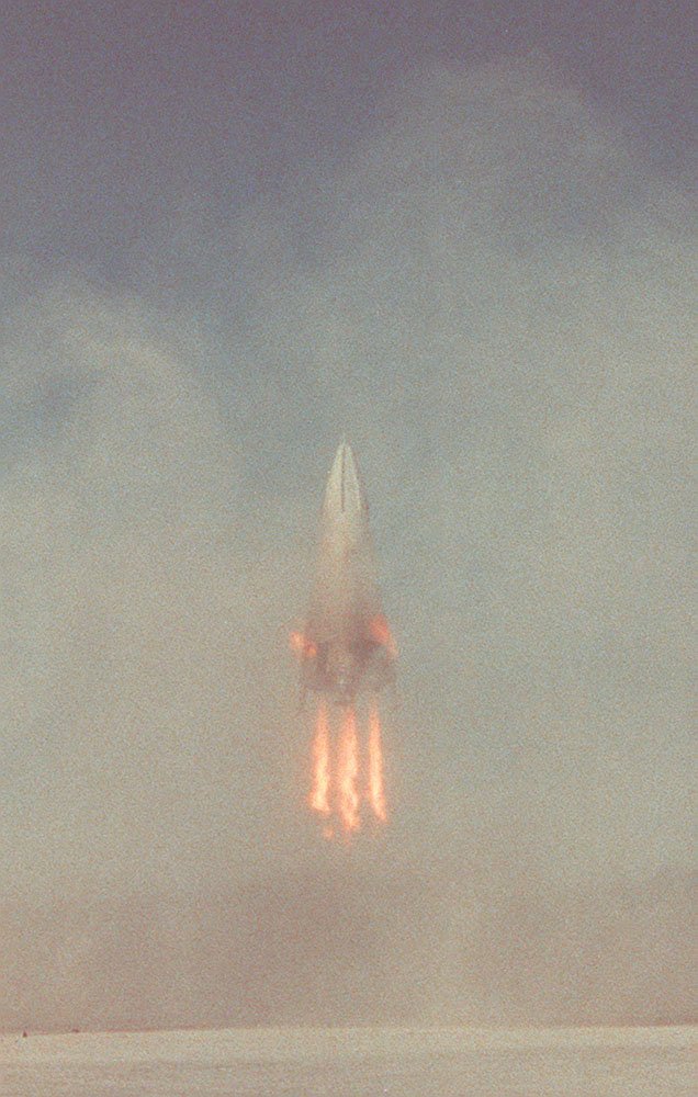 Cone-shaped rocket descending through its own dust cloud on four legs, flame beneath, desert barely visible below