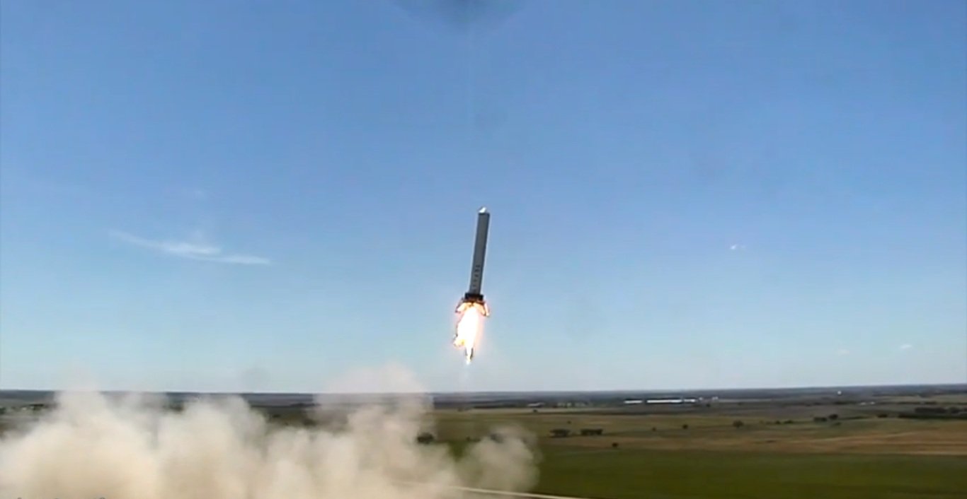 Grey cylindrical rocket hanging on a bright flame over flat Texas farmland, dust billowing from the pad