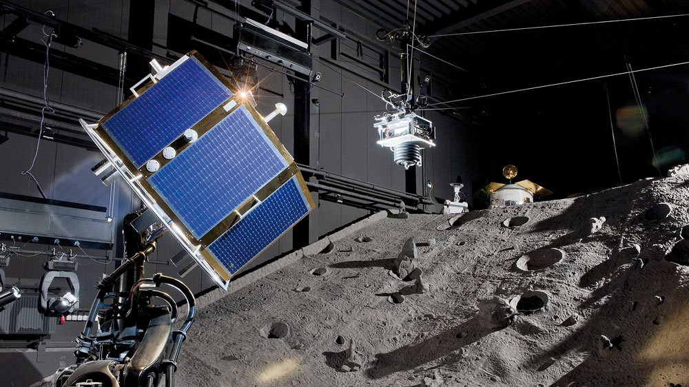 Satellite mock-up with blue solar panels hanging from cables above sculpted grey terrain