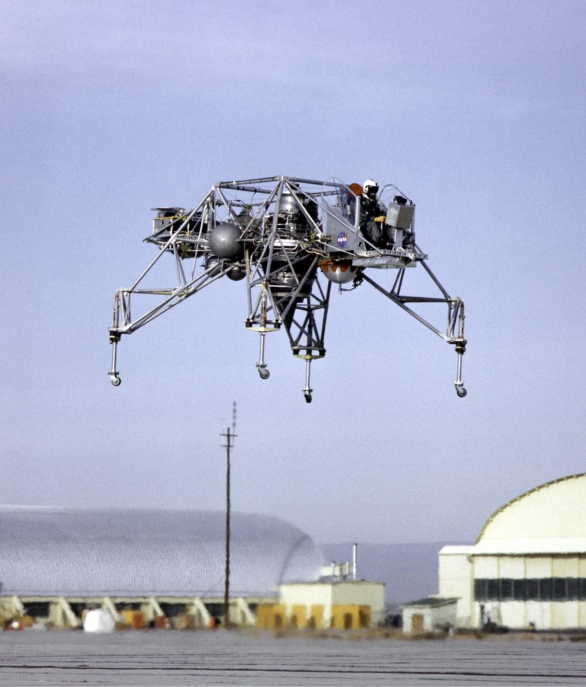Skeletal open-truss craft airborne over a desert airfield, a helmeted pilot seated in the open