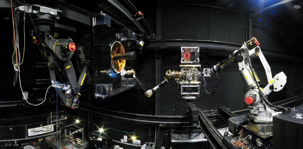 Robot arms face a mock-up with a gold adapter ring in a black-walled laboratory