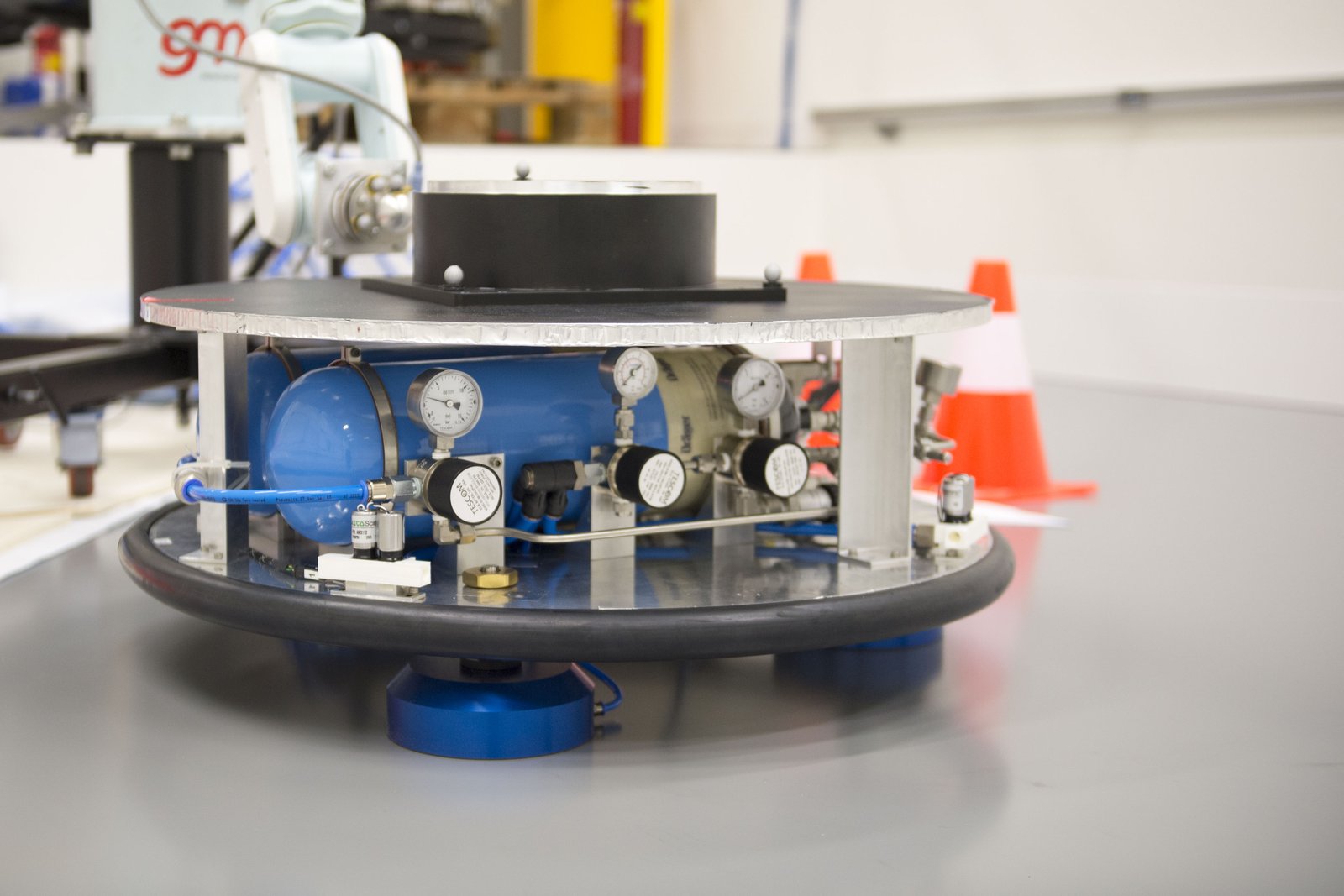 Round air-bearing platform with gas bottle and pressure gauges on a grey epoxy floor, robot arm behind