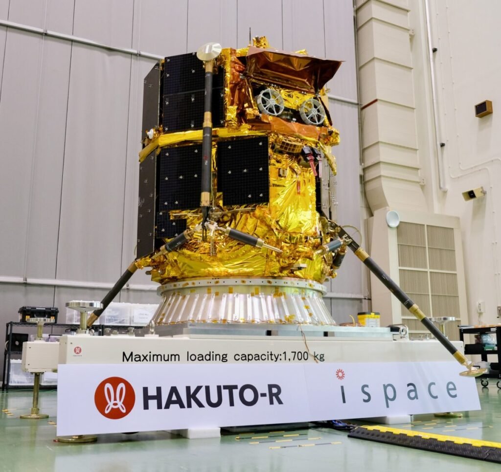 Gold-foiled lander with black solar panels and slender legs on a test stand in a cleanroom, HAKUTO-R and ispace banners below