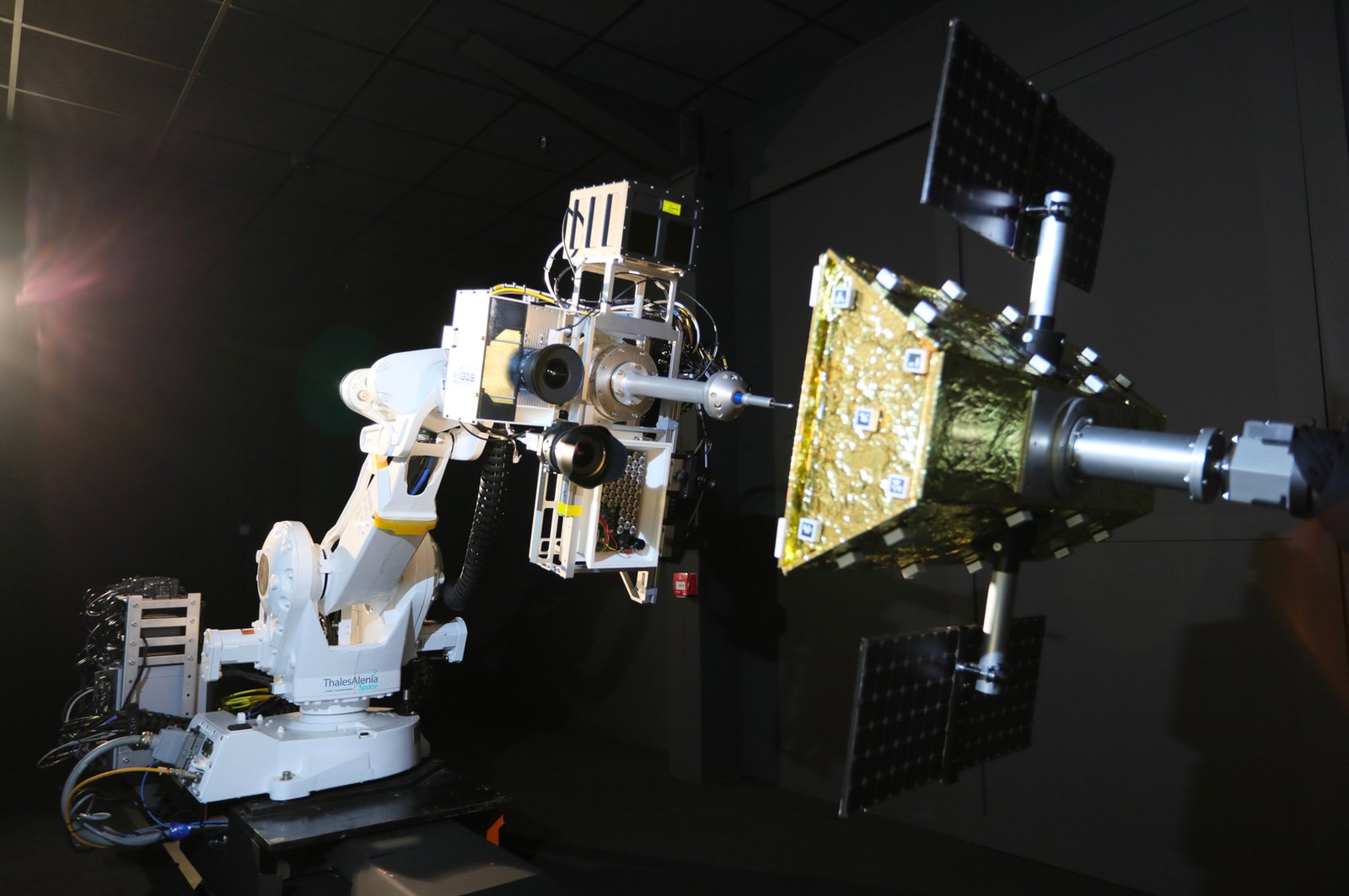 ROBY robot arm carrying a sensor head approaching a gold satellite mock-up in a dark hall