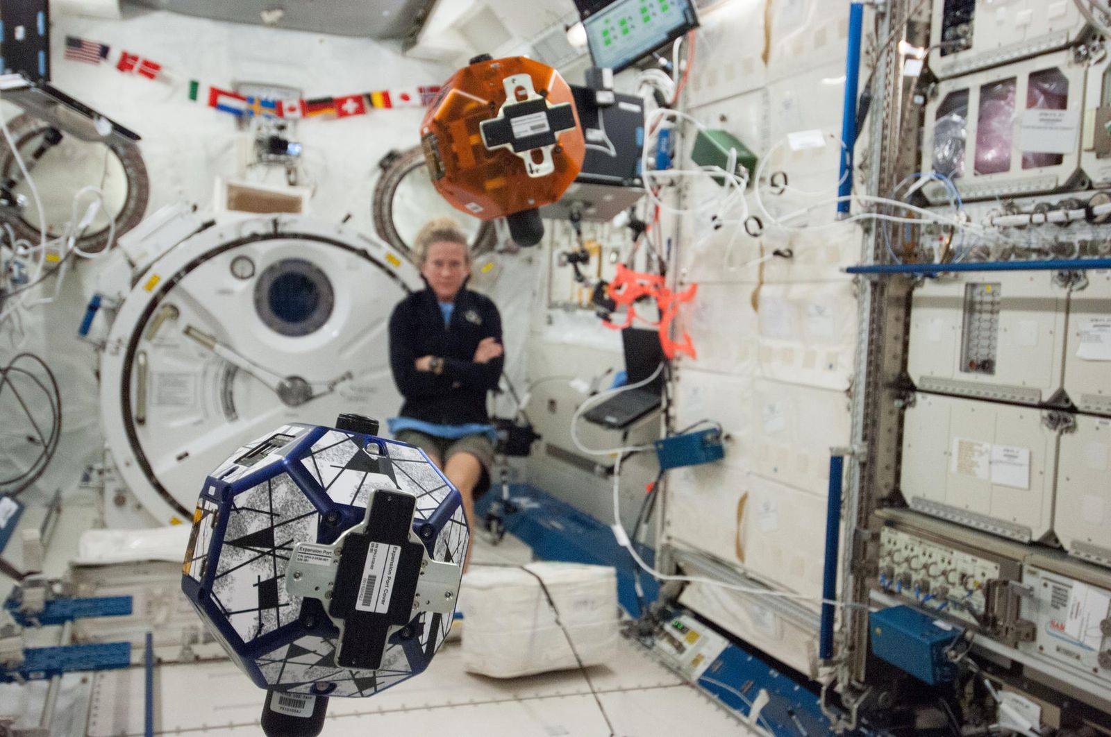 Orange and blue polyhedral satellites floating in the station cabin, an astronaut watching behind