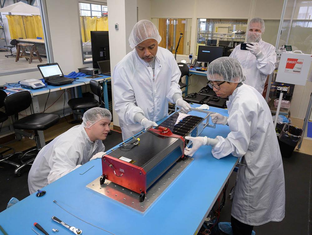 Four gowned technicians around a blue bench, handling a red chassis with a solar panel